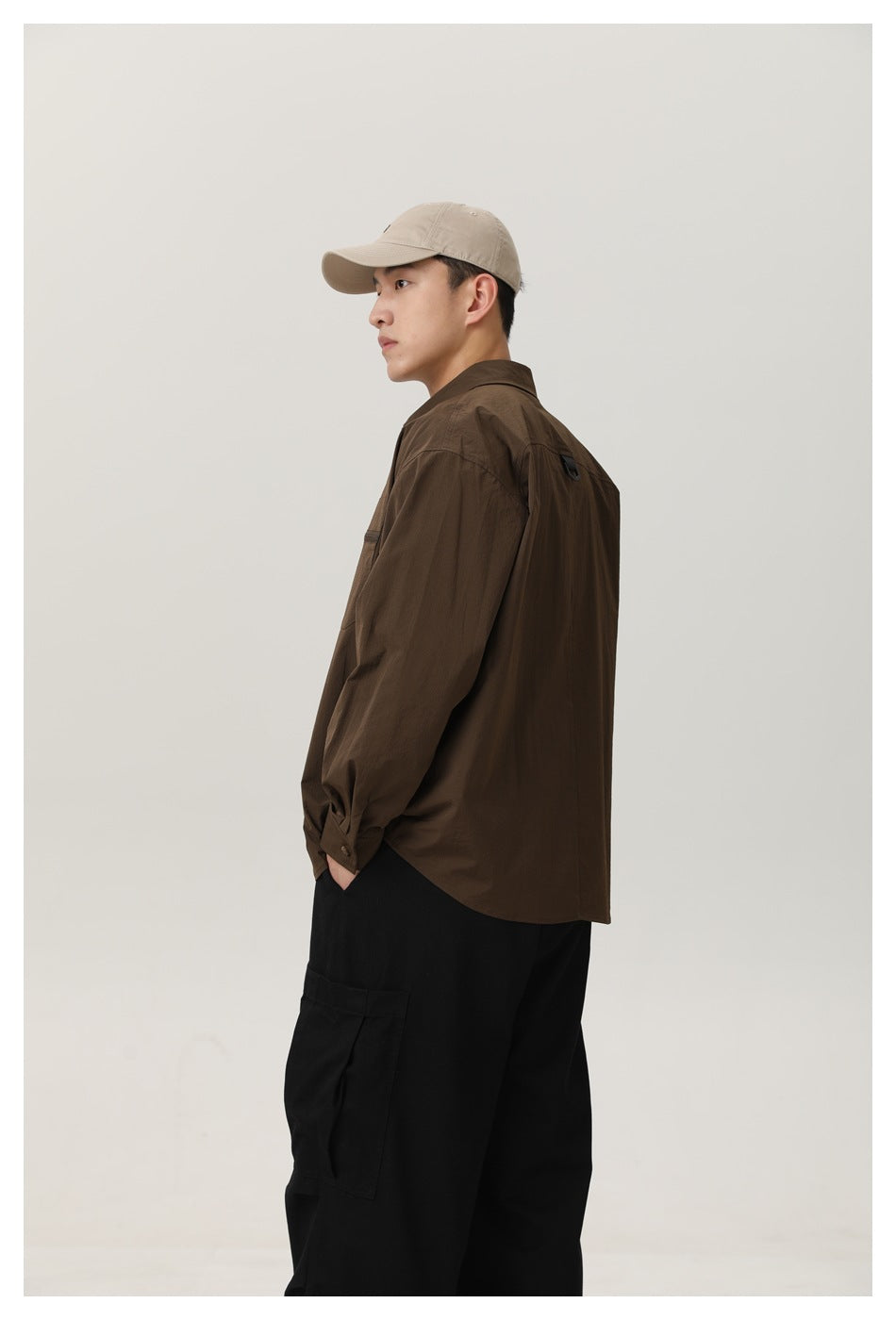 Side view of model wearing brown button-up shirt with hand in pocket, black cargo pants and beige cap