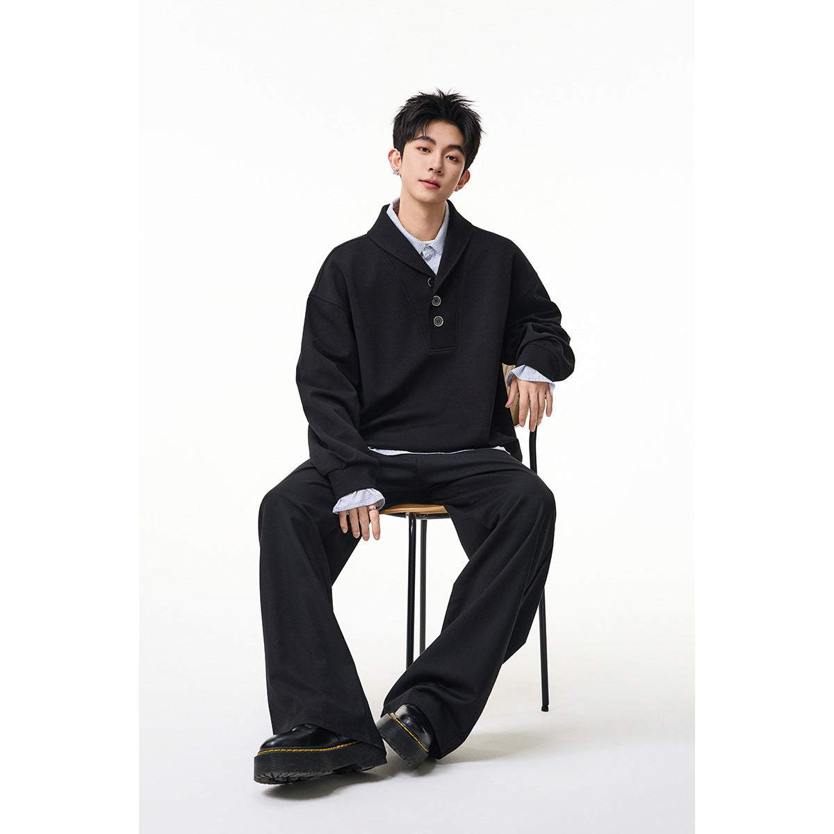 Seated model wearing a black oversized shawl-collar pullover with a three-button placket and pale blue shirt.