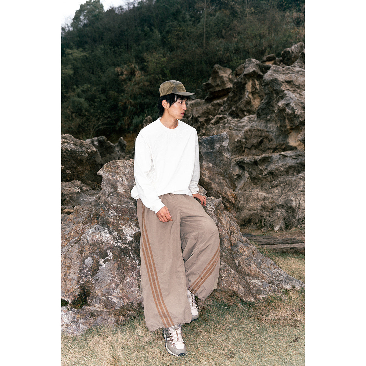 Model seated on rocks wearing a white long-sleeve shirt and taupe wide-leg pants with side stripes.