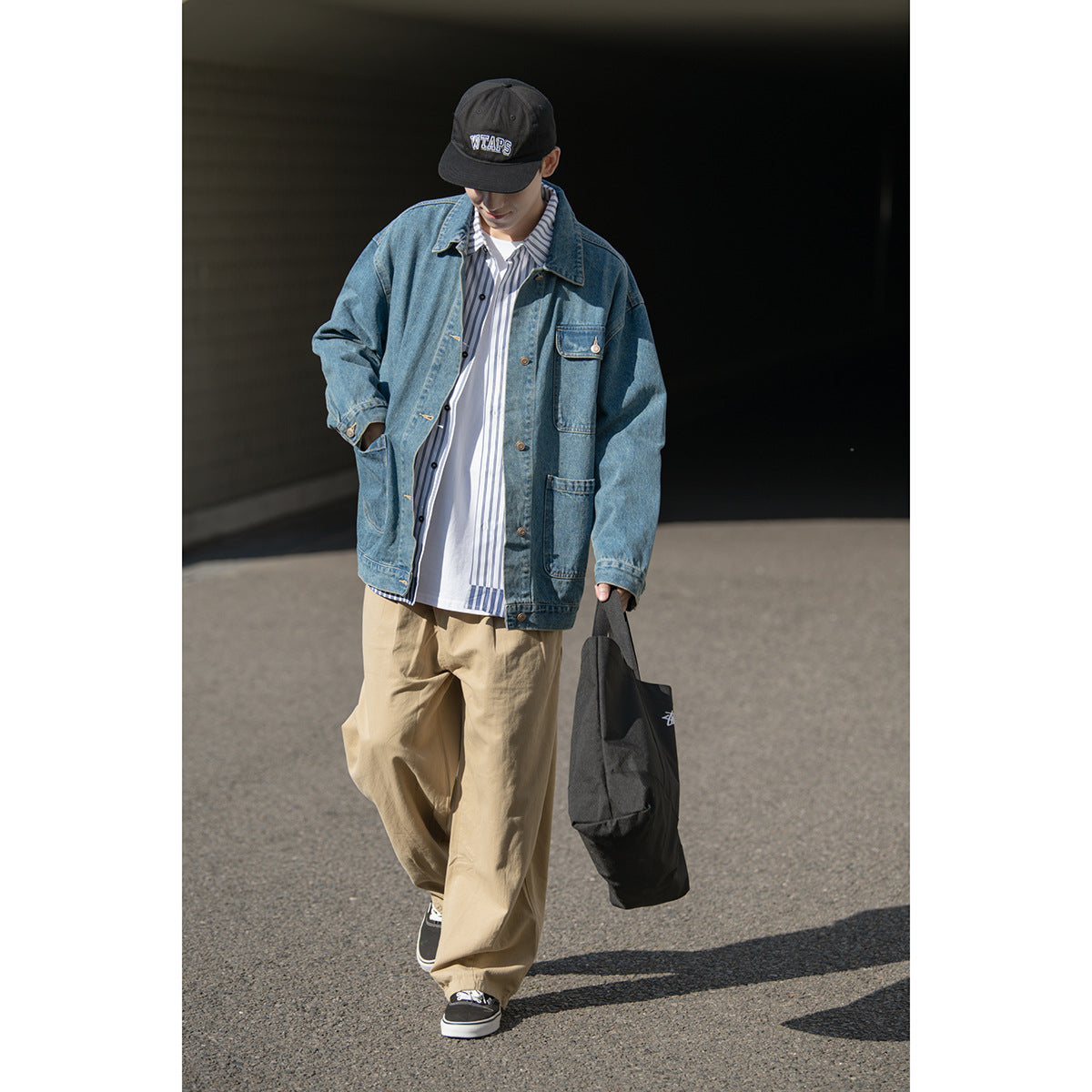 Model walking in a blue denim jacket with patch pockets, a striped shirt, and loose tan trousers.