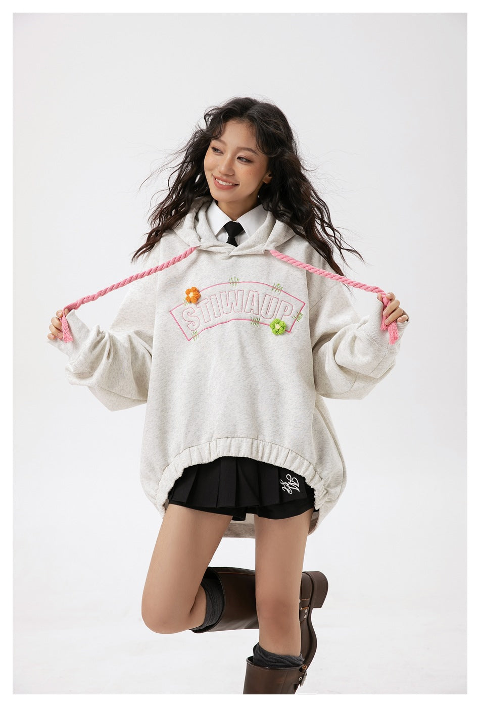 Model wearing an oversized light gray hoodie with pink rope drawstrings, floral embroidery and arched lettering.