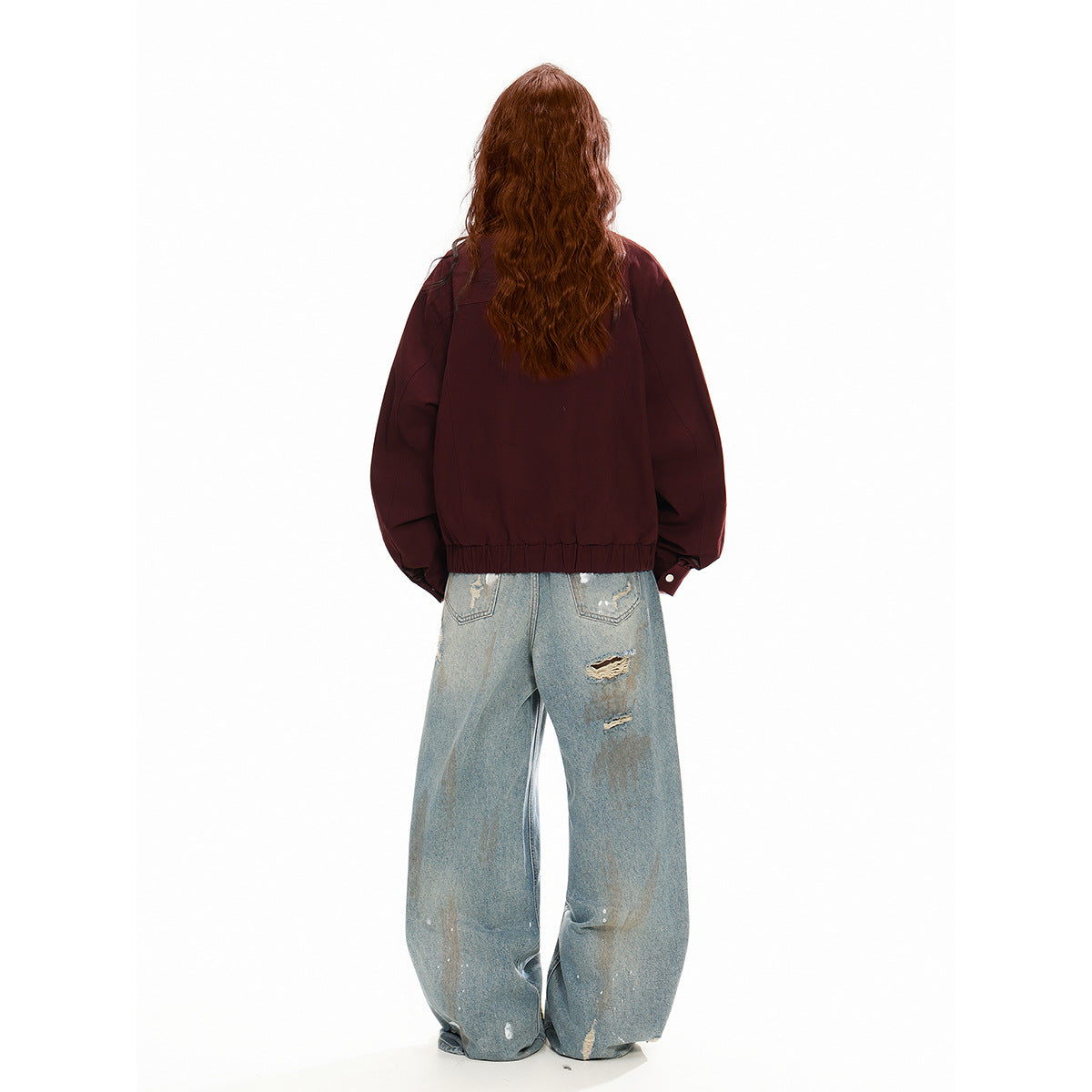 Rear view of a model wearing a burgundy jacket with an elastic hem and loose, distressed light-wash jeans.