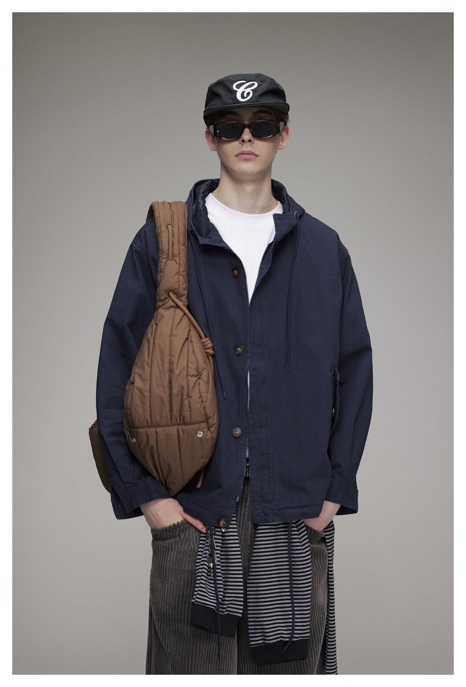 Model wearing a navy hooded button-front jacket over a white T-shirt, with a brown shoulder bag.