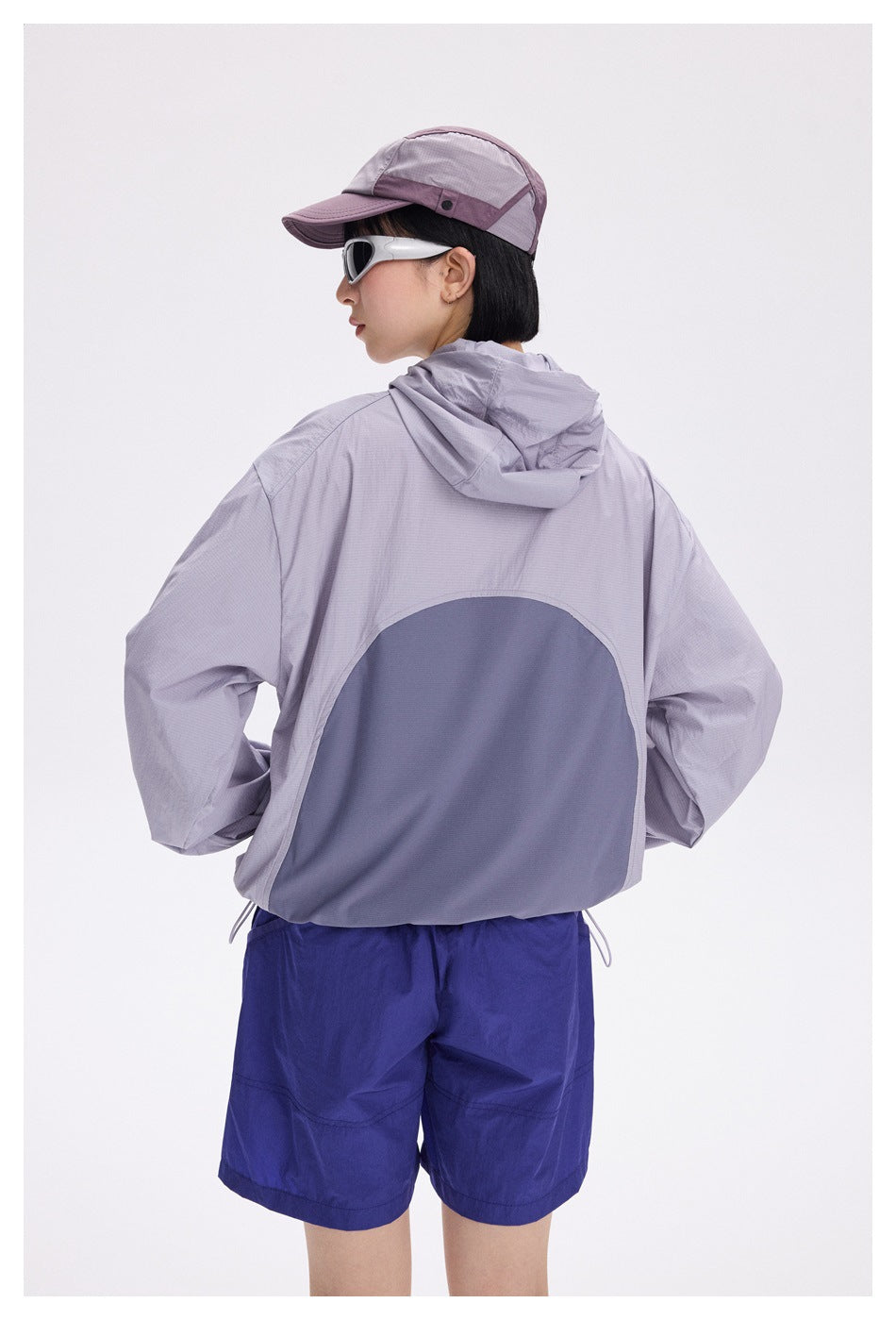 Rear view of a model in a light lavender hooded jacket with a dark curved back panel, royal blue shorts and a cap.