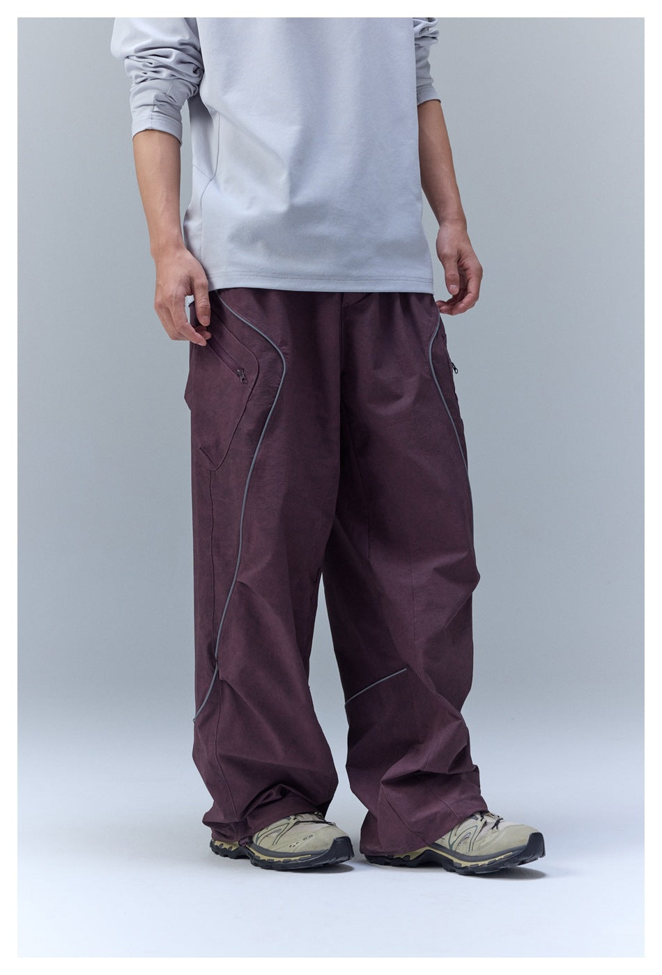 Cropped model wearing loose plum trousers with curved contrast piping and a large side pocket.