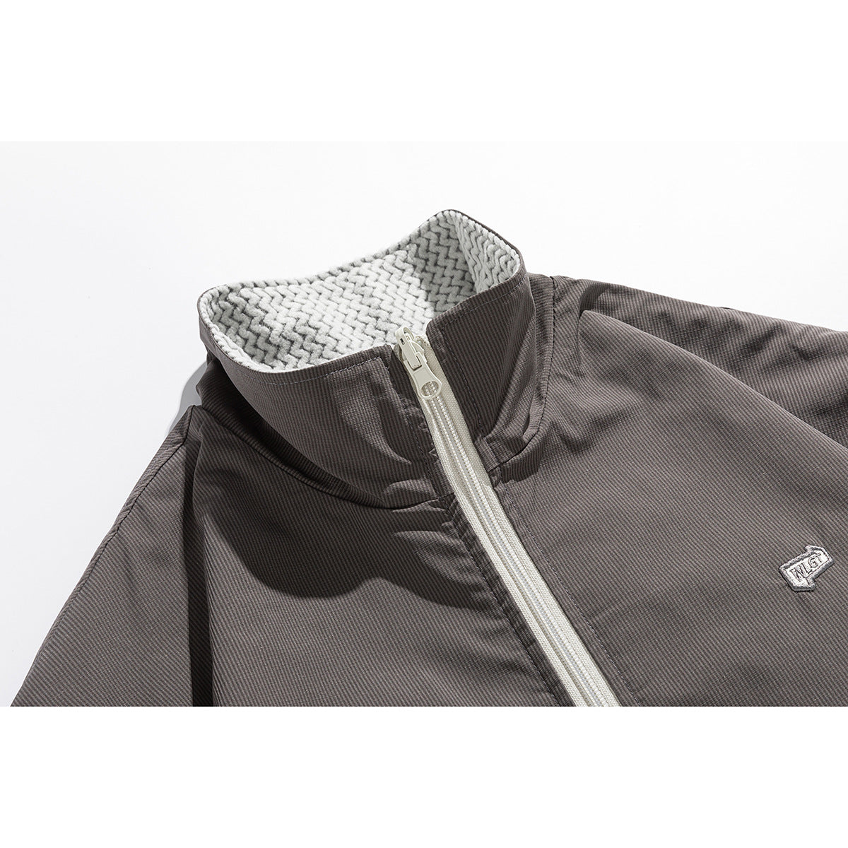 Close-up of a charcoal jacket's raised collar, patterned lining, pale zipper and small embroidered chest emblem.