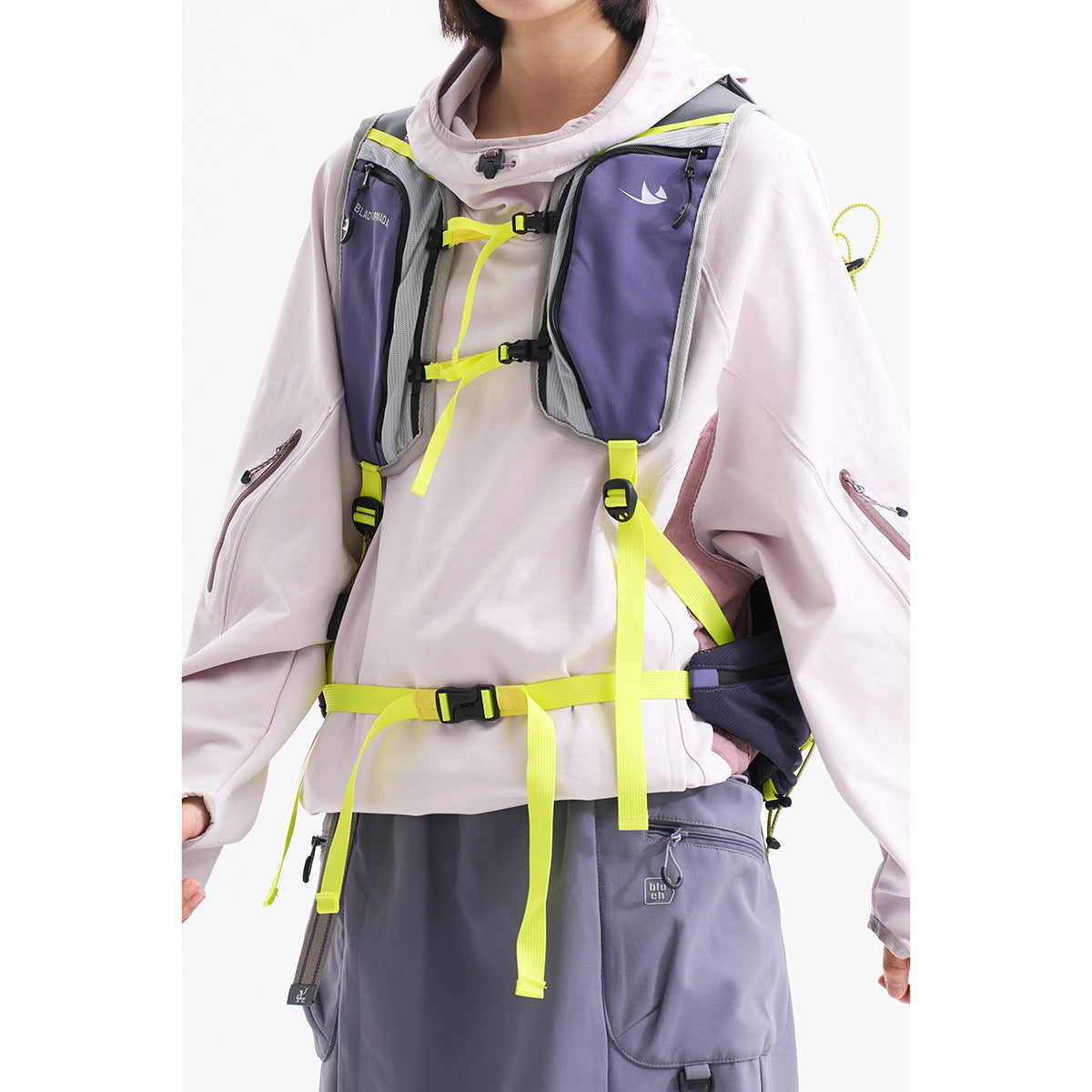 Close view of a model wearing a lavender-gray chest pack with neon yellow straps over a pale hooded jacket.