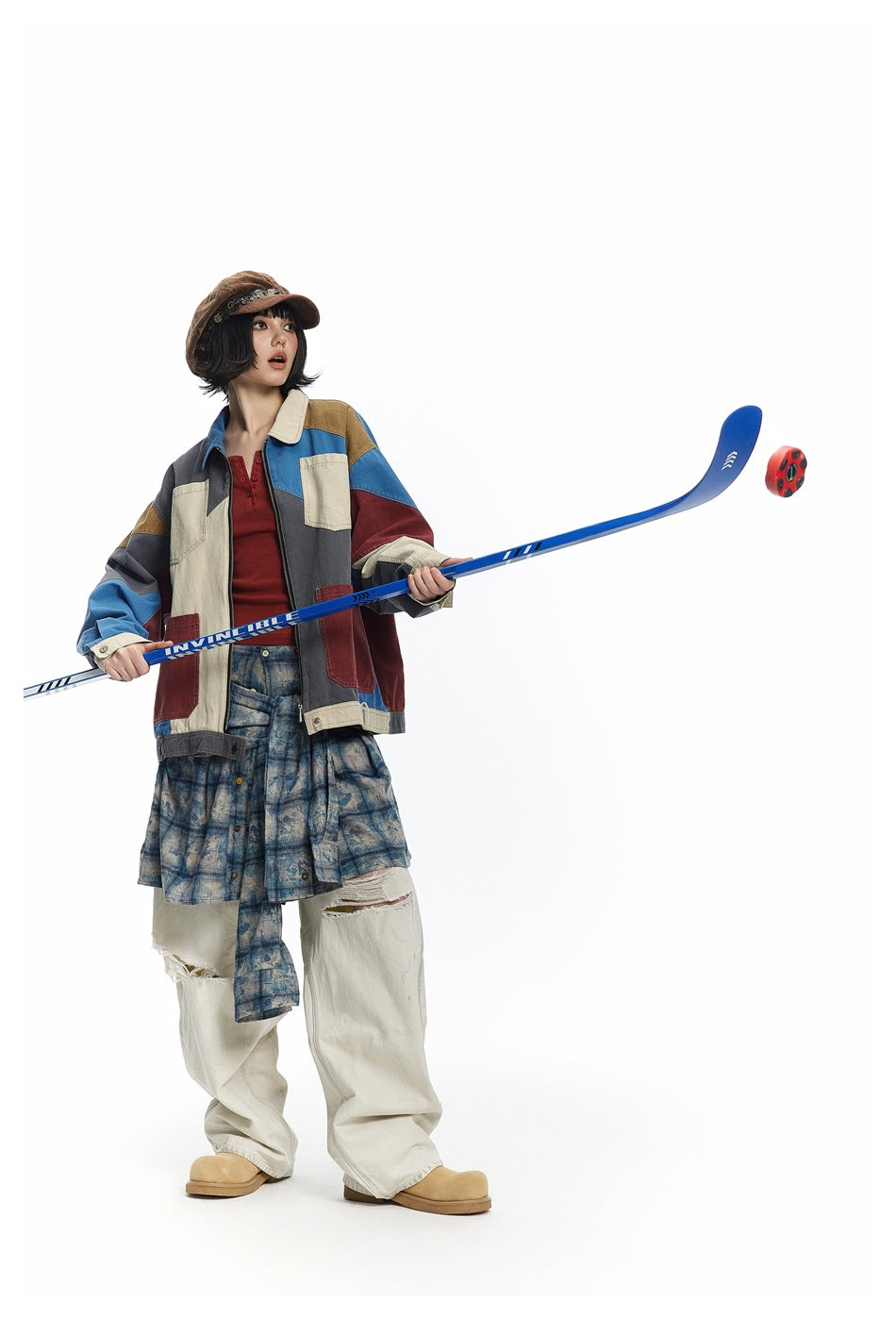 Model wearing a multicolor patchwork jacket and blue plaid skirt while holding a blue hockey stick.