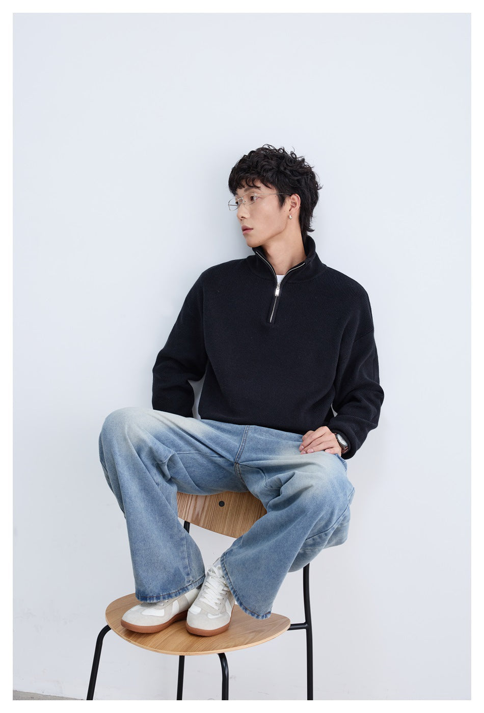 Seated model wearing a black quarter-zip sweater and loose blue jeans against a pale studio background.
