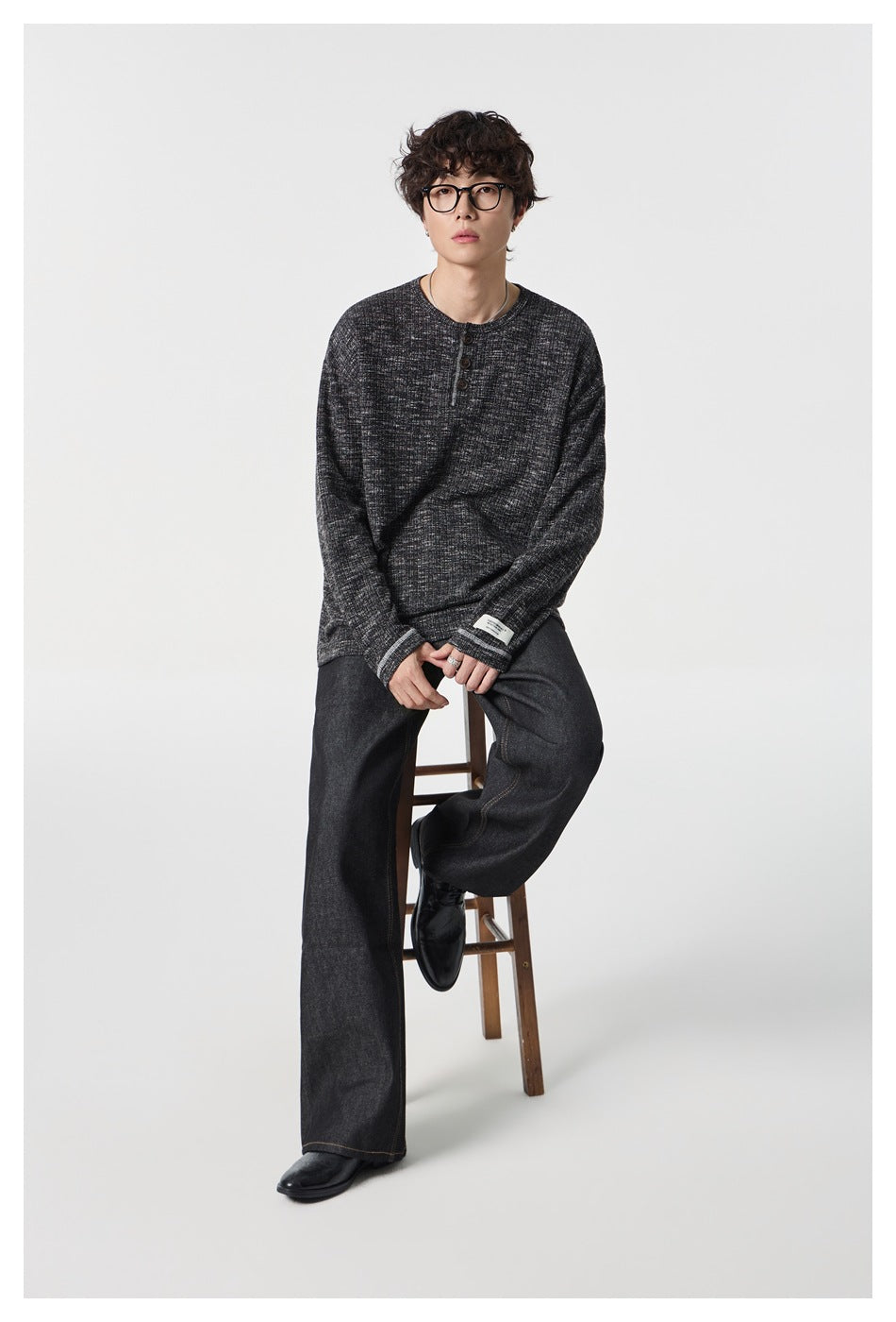 Model seated on a stool wearing a charcoal heathered long-sleeve henley and dark wide-leg jeans.
