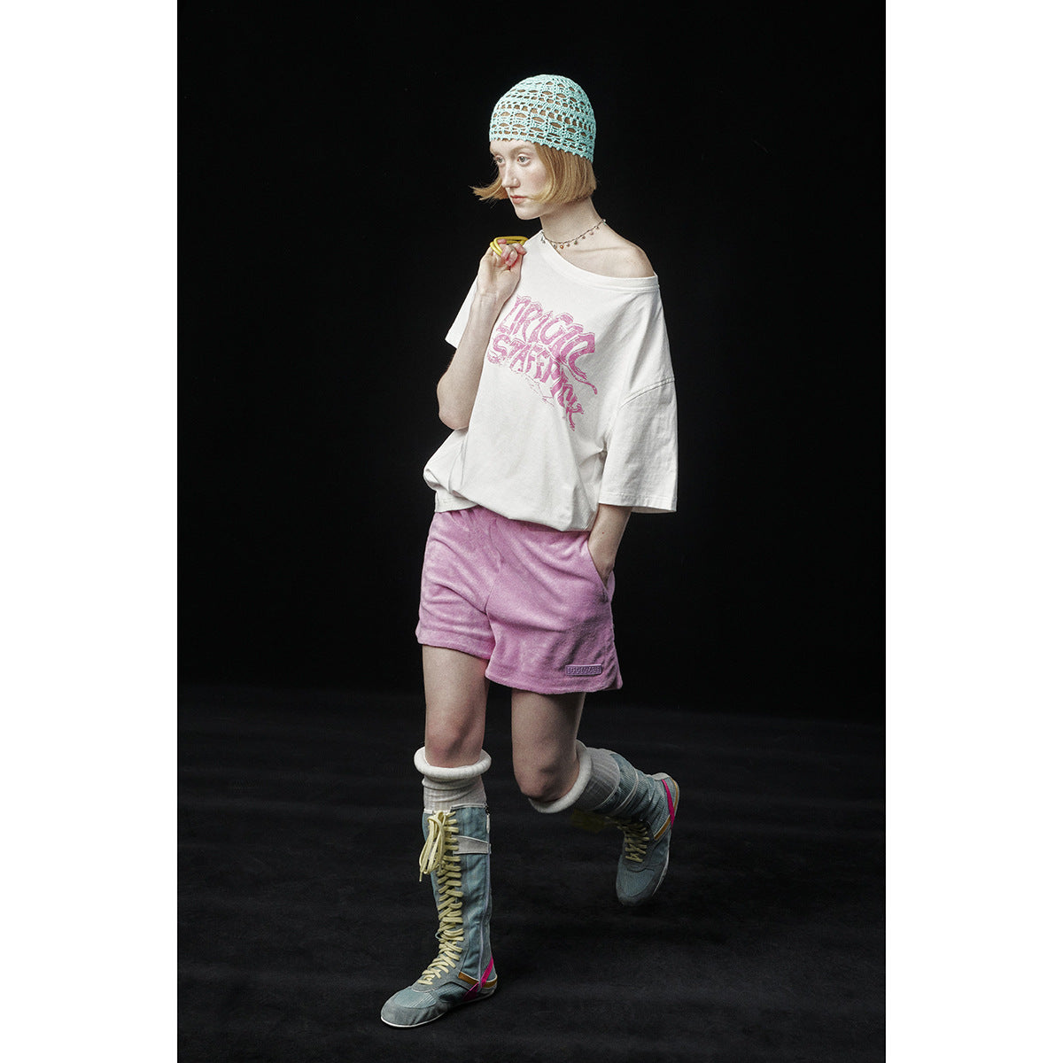 Model wearing an oversized white graphic T-shirt, pink shorts, crocheted cap and tall lace-up boots.
