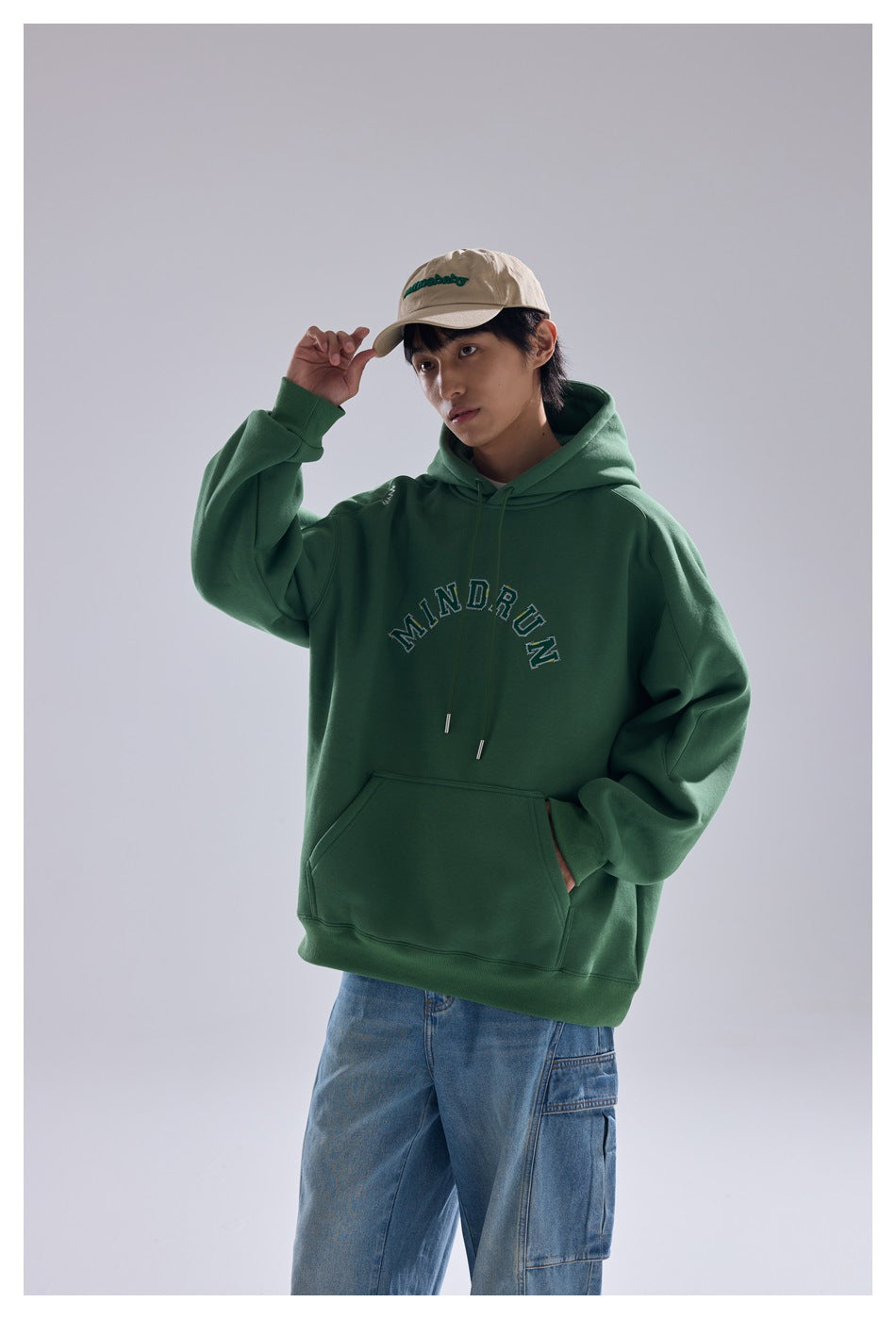Model wearing a green MINDRUN hoodie with a beige cap and light-wash jeans, hand raised to the cap brim
