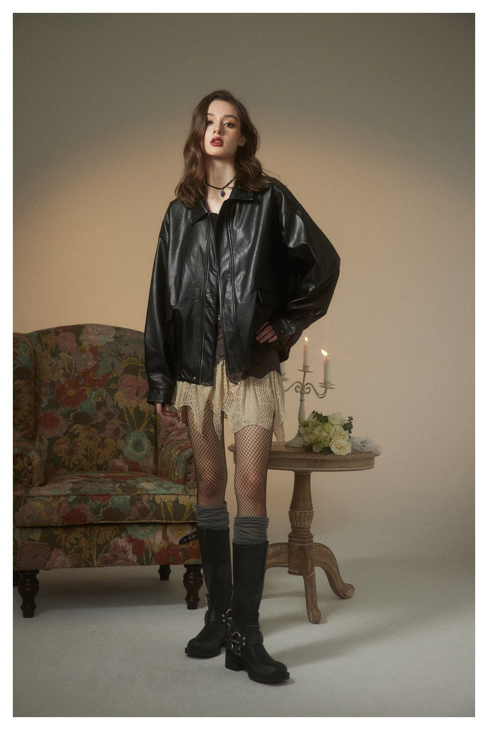 Model wearing an oversized glossy black zip-front jacket with a pointed collar, styled with a cream lace skirt and boots.
