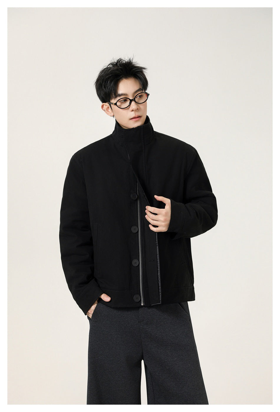 Model wearing a black high-collar jacket with a front zipper and buttons, paired with charcoal wide-leg trousers.