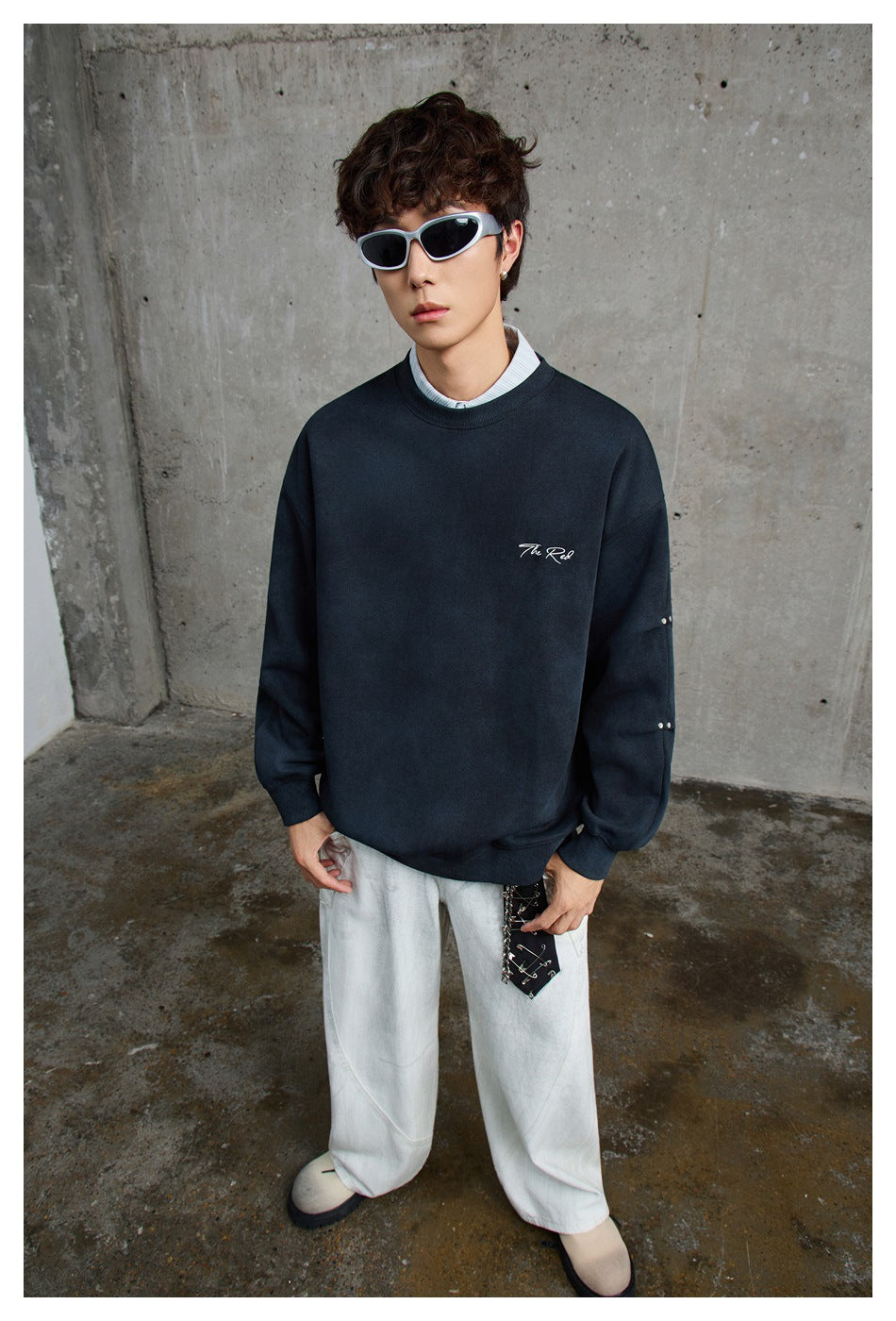 Model wearing a dark navy crewneck sweatshirt with small white chest script and sleeve studs, styled with white trousers.