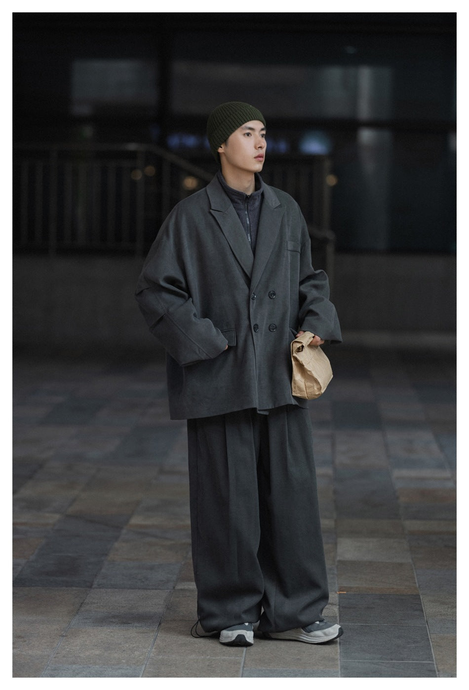 Model wears a dark gray oversized double-breasted coat over a matching outfit and olive knit beanie.