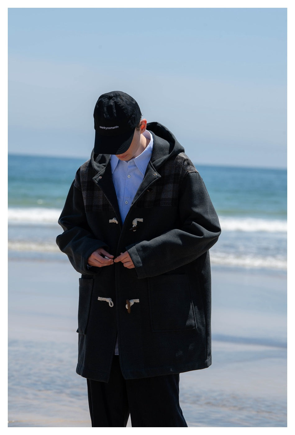 Person fastening a dark hooded duffle coat with plaid shoulder panels and large front pockets.