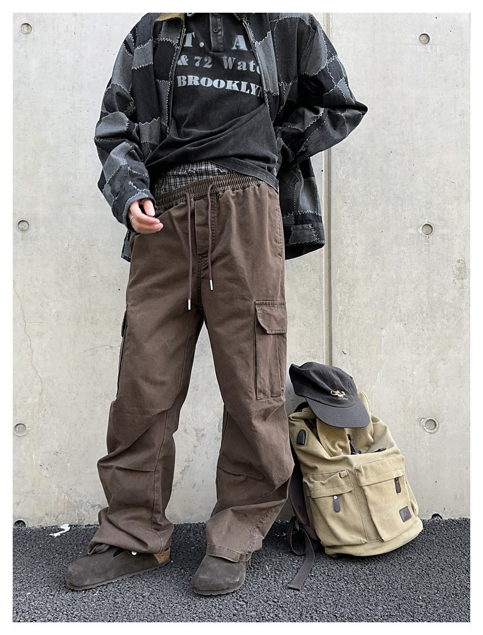 Person wearing brown wide-leg cargo pants with a patchwork denim jacket, standing by a concrete wall next to a backpack