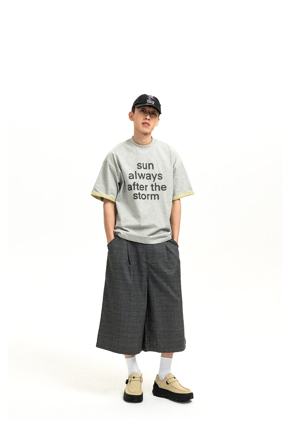 Model standing with hands in pockets wearing dark gray plaid wide-leg shorts, gray graphic T-shirt and beige shoes