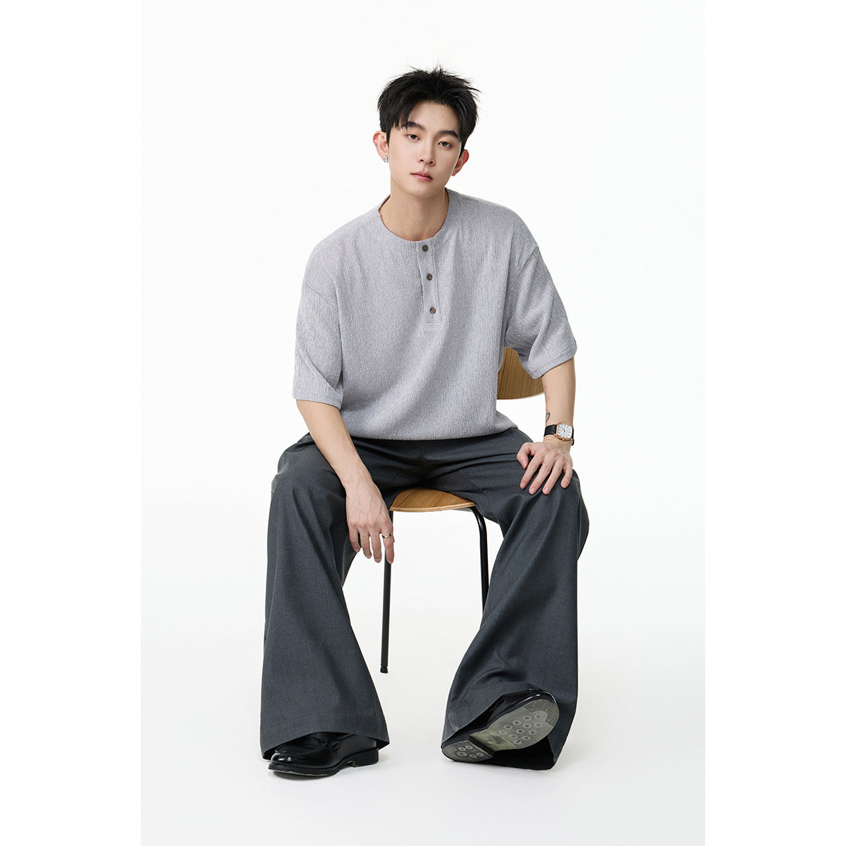 Seated model wearing a light gray short-sleeve henley shirt and dark wide-leg trousers.