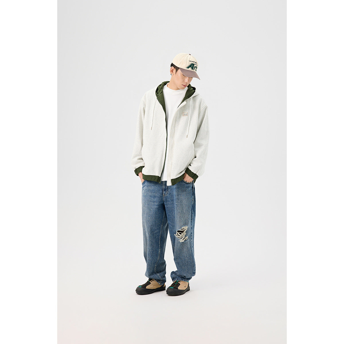 Model wearing a light gray hooded zip jacket with dark green lining and trim, styled with loose blue jeans.