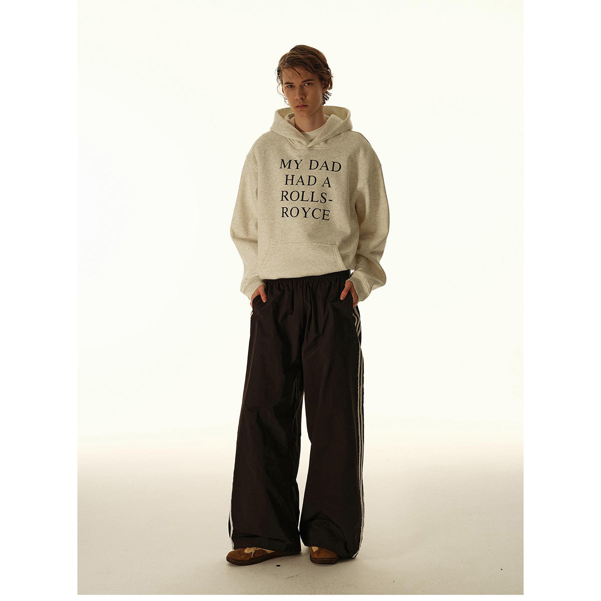 Model wearing a cream hoodie printed “MY DAD HAD A ROLLS-ROYCE” with wide black striped pants.