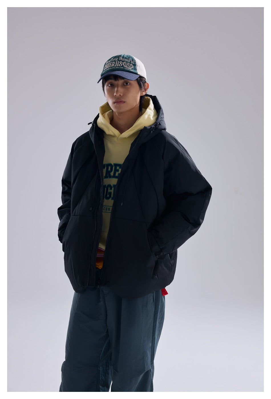 Model in an open black hooded puffer jacket over a yellow hoodie, hands in pockets, close-up three-quarter view