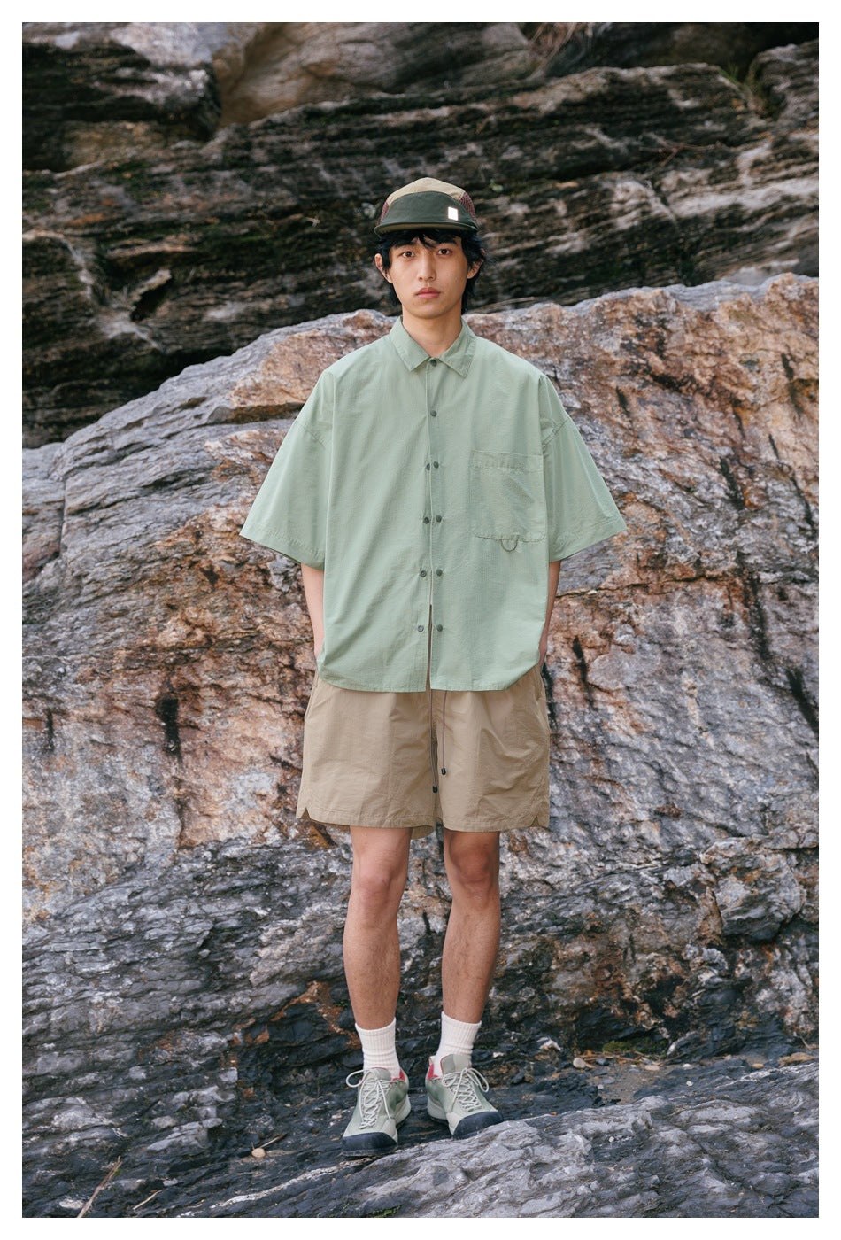 Model wearing a pale sage-green short-sleeve button-up shirt and tan shorts, standing in front of rocks.