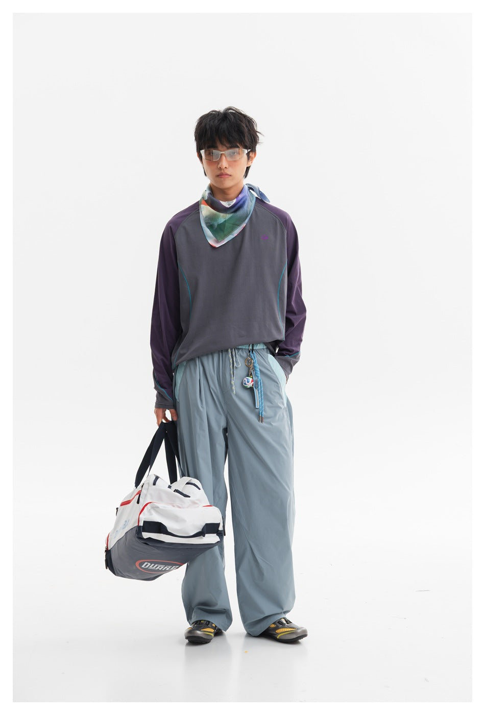 Front view of a model wearing a charcoal top with purple sleeves and teal piping, loose blue-gray trousers, and a patterned