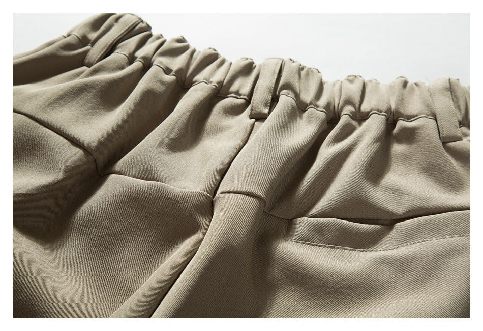 Close-up of taupe trousers showing a gathered waistband, belt loops, pleats and front pocket openings.
