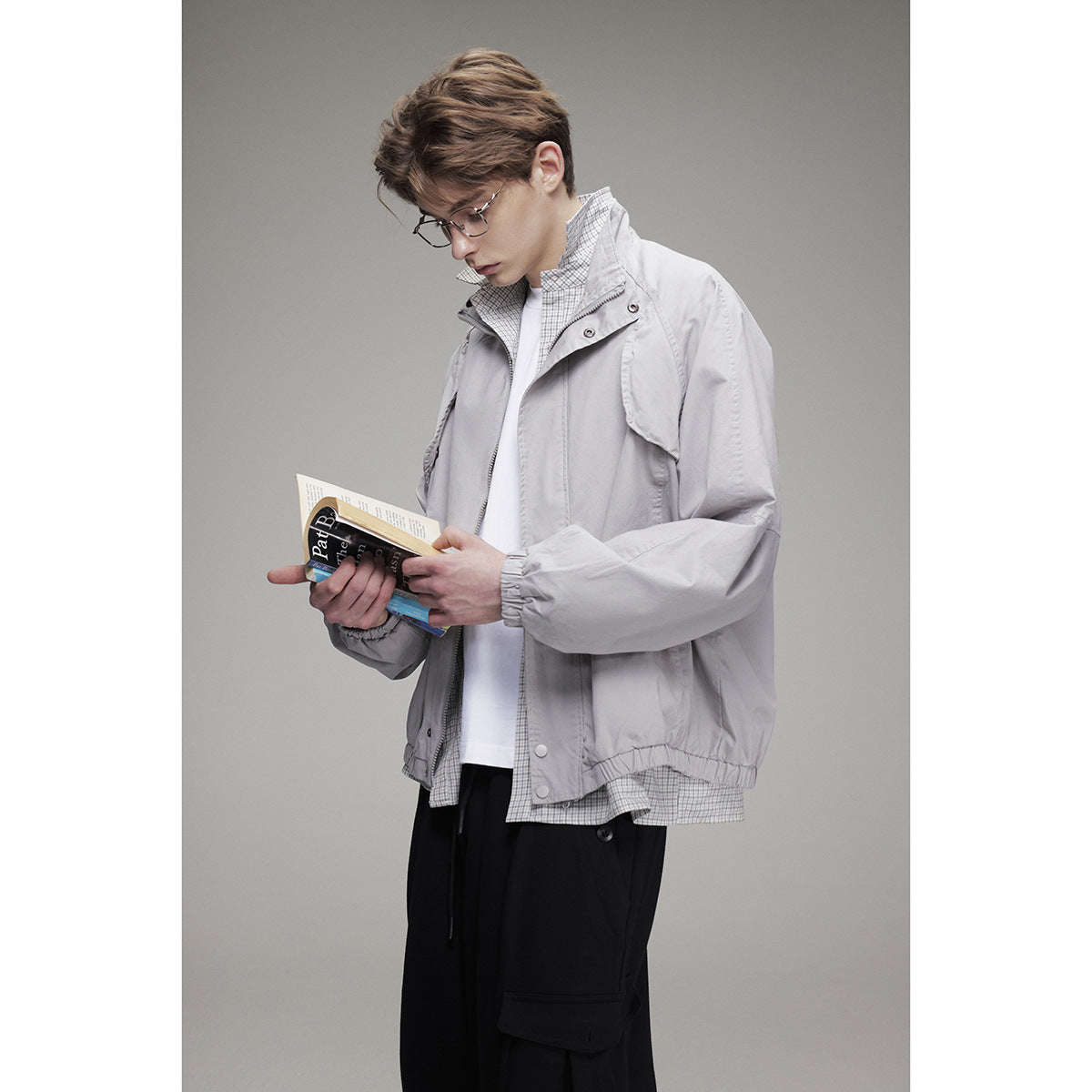 Model wearing a light gray jacket with a raised collar and flap pockets, holding an open book.