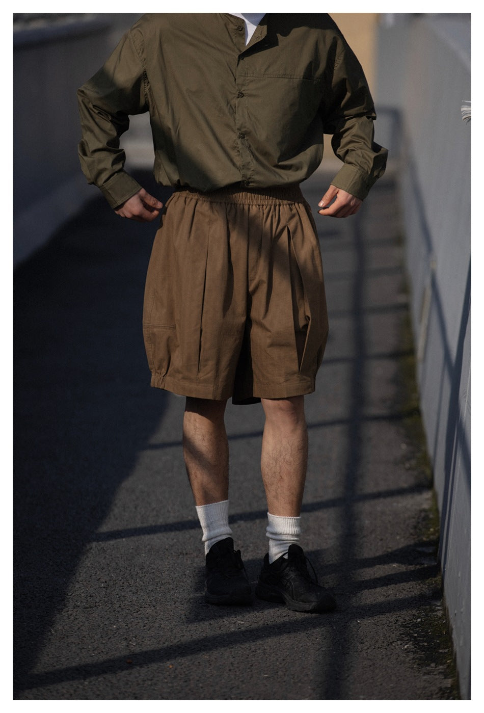 Model wearing brown loose shorts with an elastic waist, an olive button-up shirt, white socks and black sneakers.