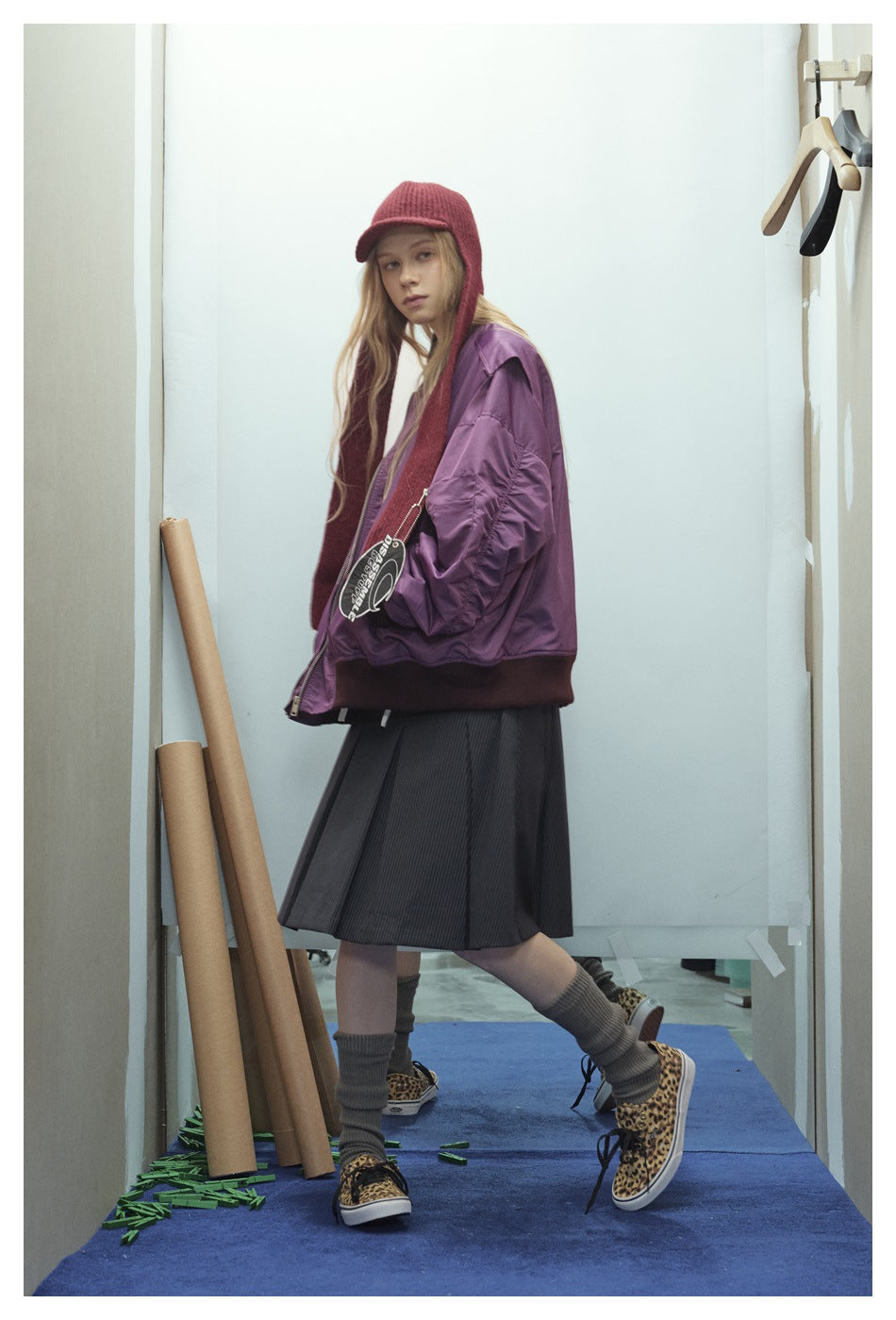 Model wearing an oversized purple bomber jacket with a red knit hat, gray pleated skirt and leopard sneakers