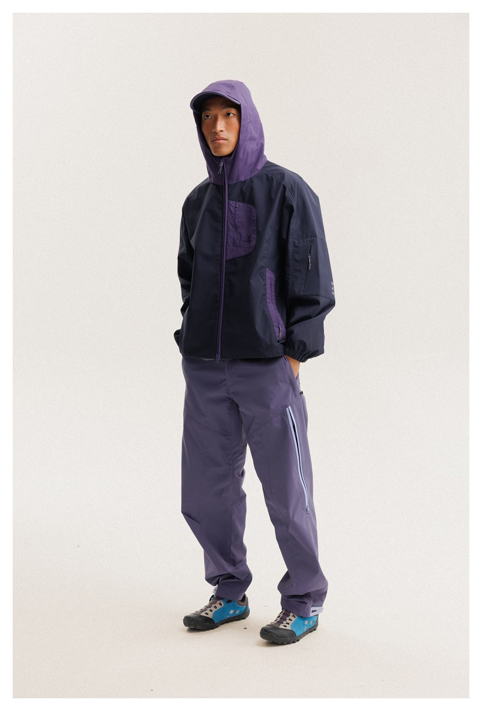 Three-quarter view of a model in a navy jacket with purple hood and purple pants, standing with hands in pockets.
