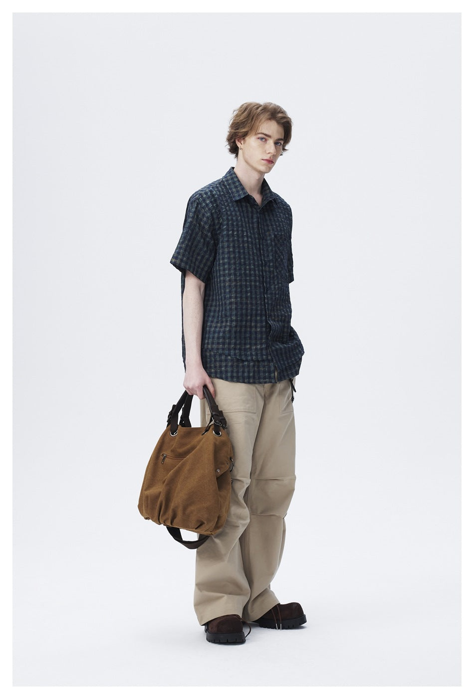 Model in dark check short-sleeve shirt with beige cargo pants, turned at an angle holding a brown bag
