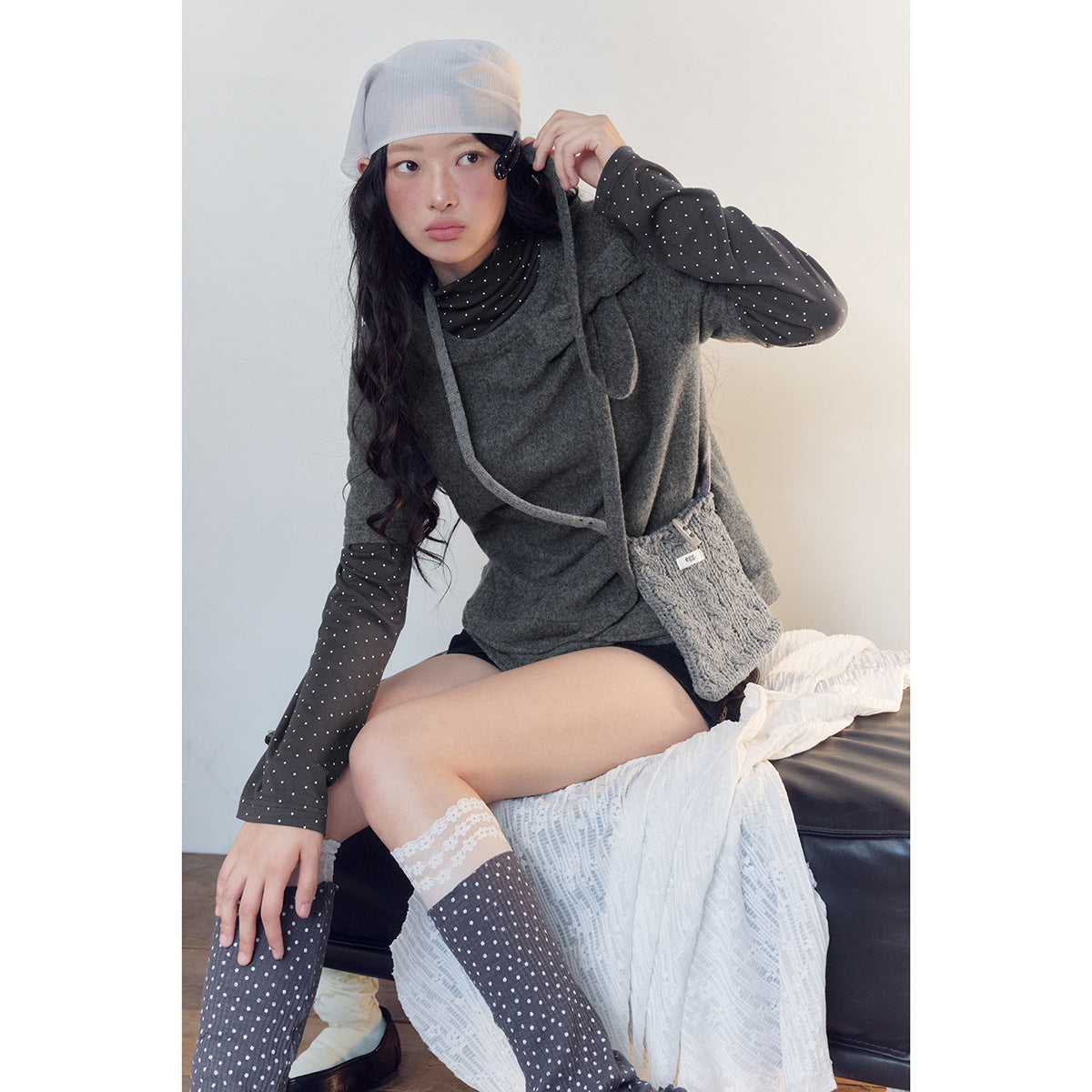 Model wearing a charcoal gray short-sleeve top with a bow detail over a black polka-dot layer, with a knitted crossbody bag.