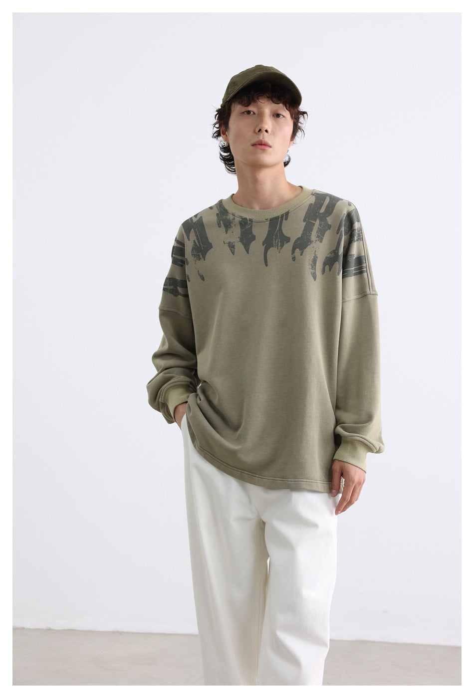 Model wearing a khaki sweatshirt with dark abstract shoulder graphics and white pants, shown from the front.