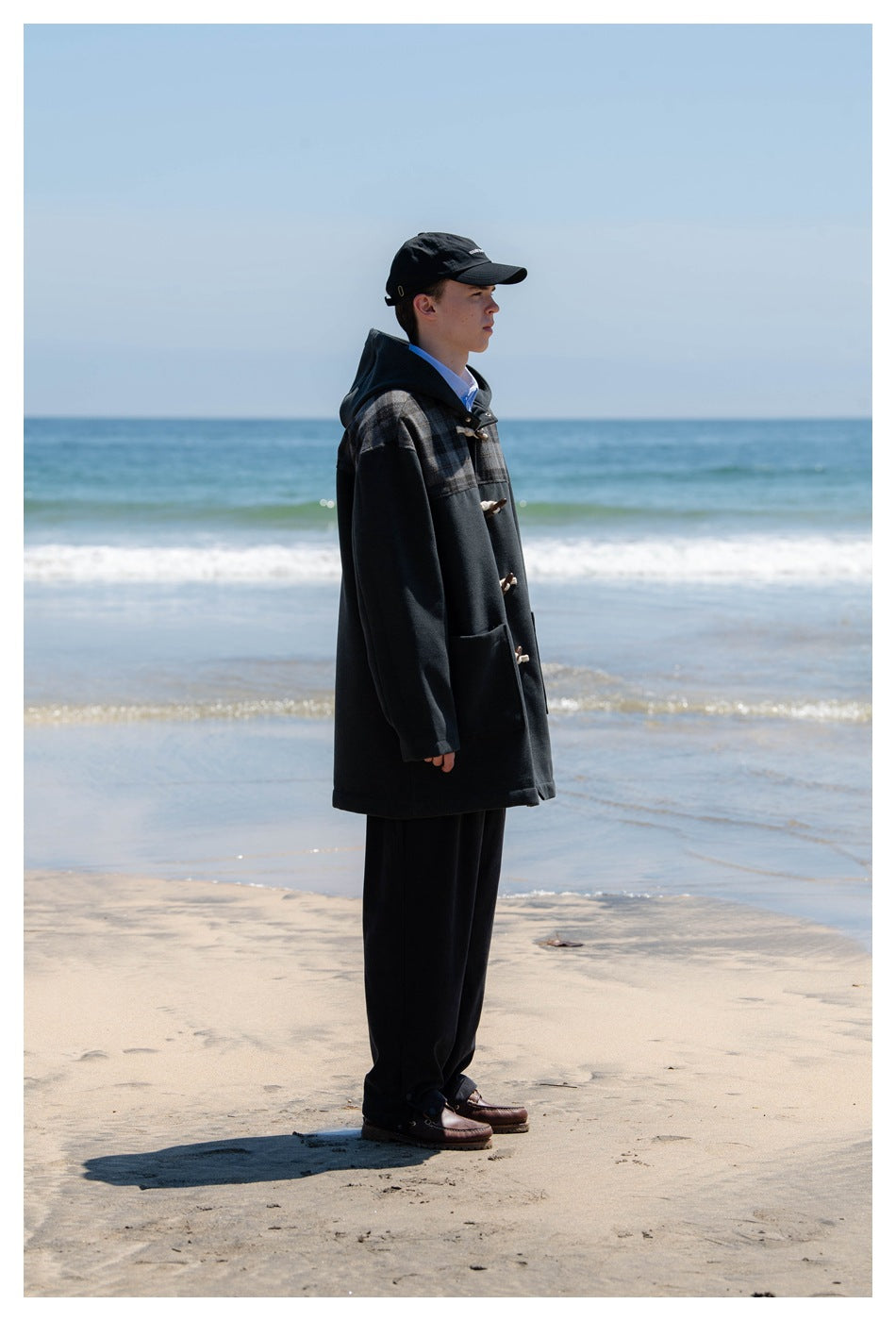 Side view of a model wearing a charcoal hooded coat with plaid shoulders and toggle closures on a beach.
