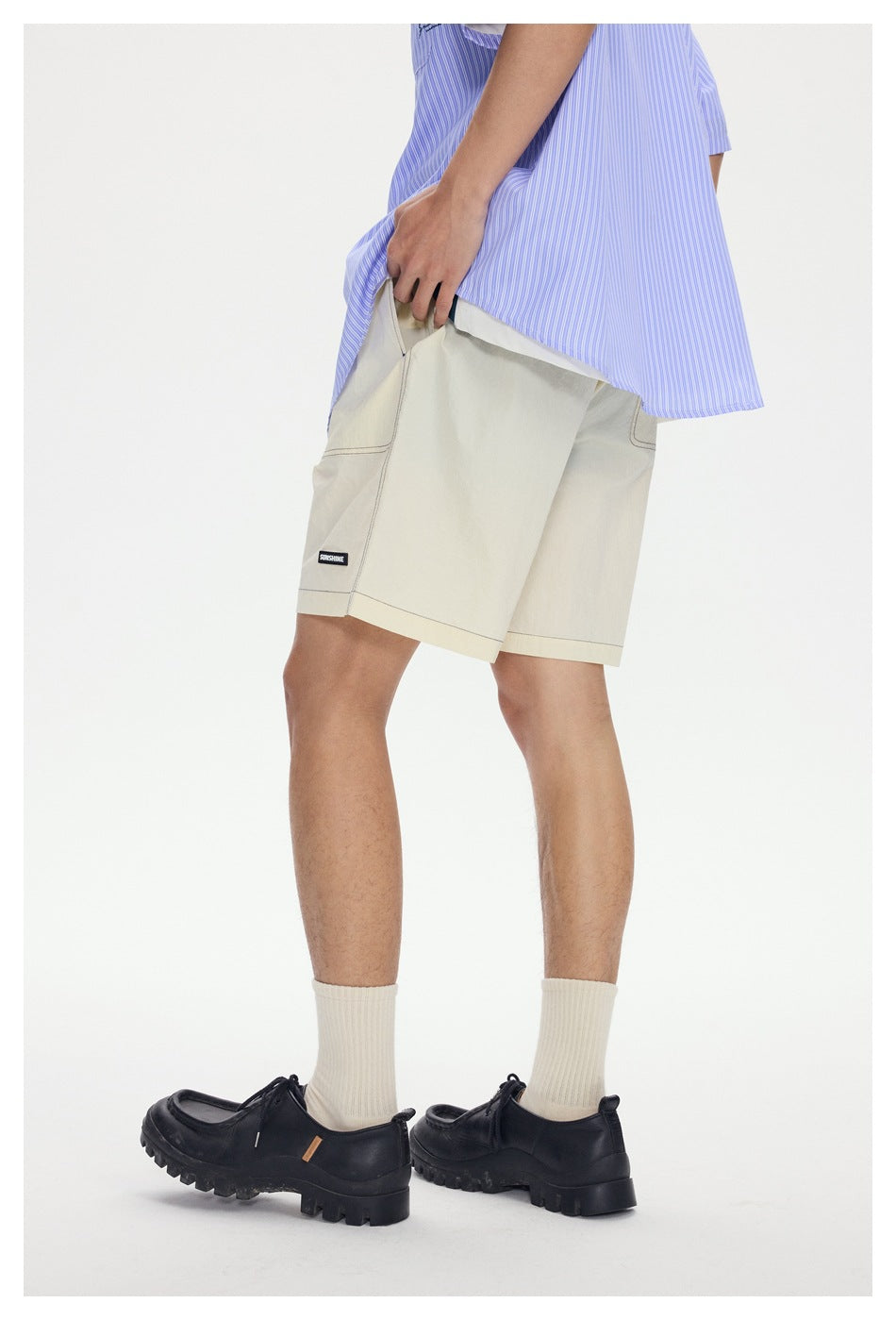 Angled side view of a model wearing cream knee-length shorts with a small black label and a blue striped shirt.