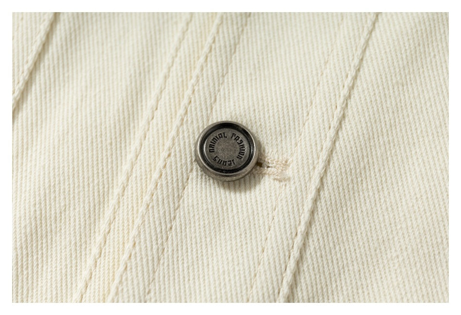 Close-up of a dark metal button on cream twill fabric, with stitched seams.