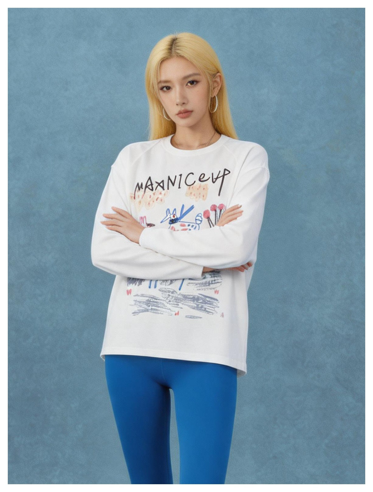 Model with long blonde hair wearing a white graphic long-sleeve tee with blue leggings, arms crossed, against a blue backdrop