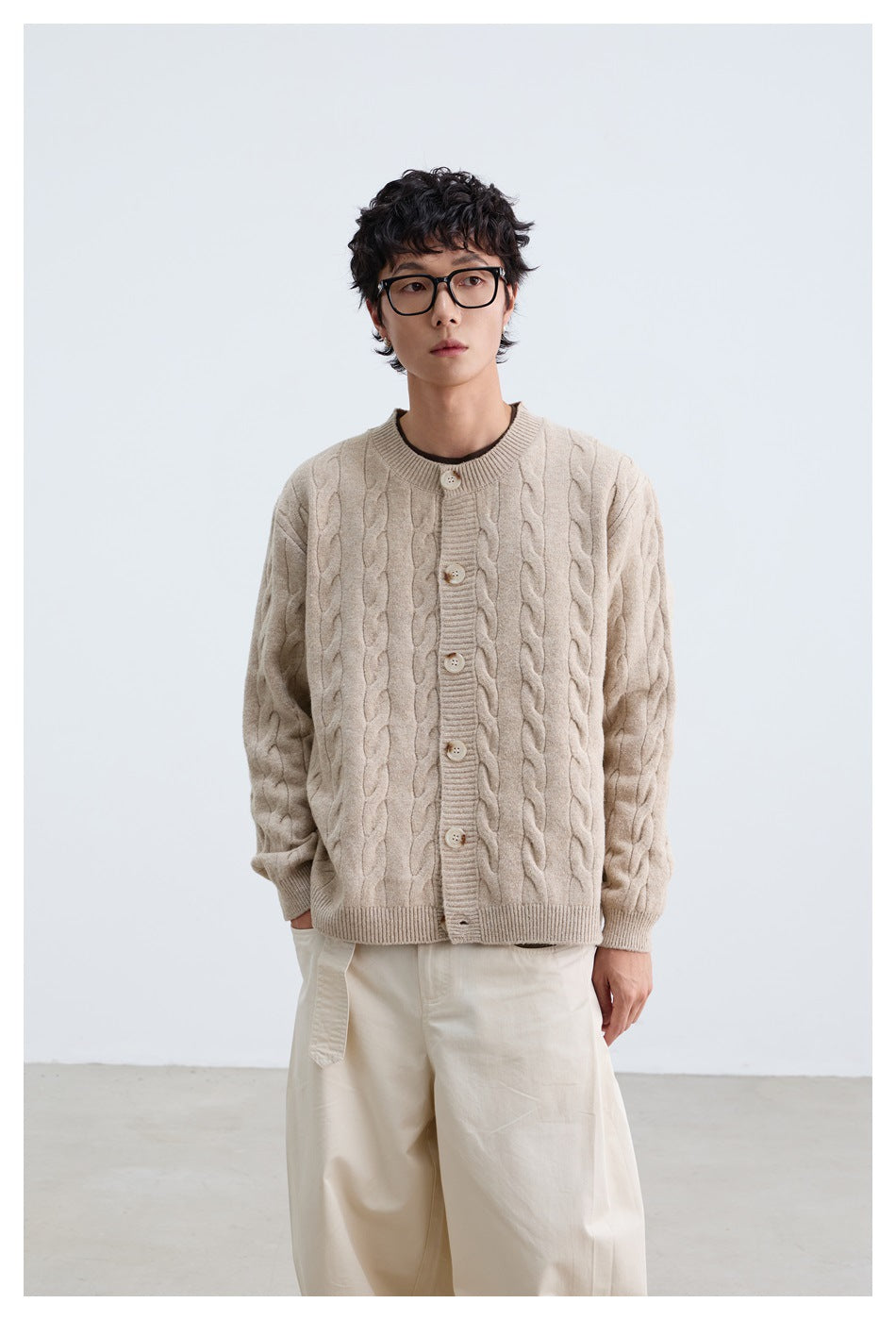 Standing model wearing a light beige cable-knit button cardigan with loose white trousers.