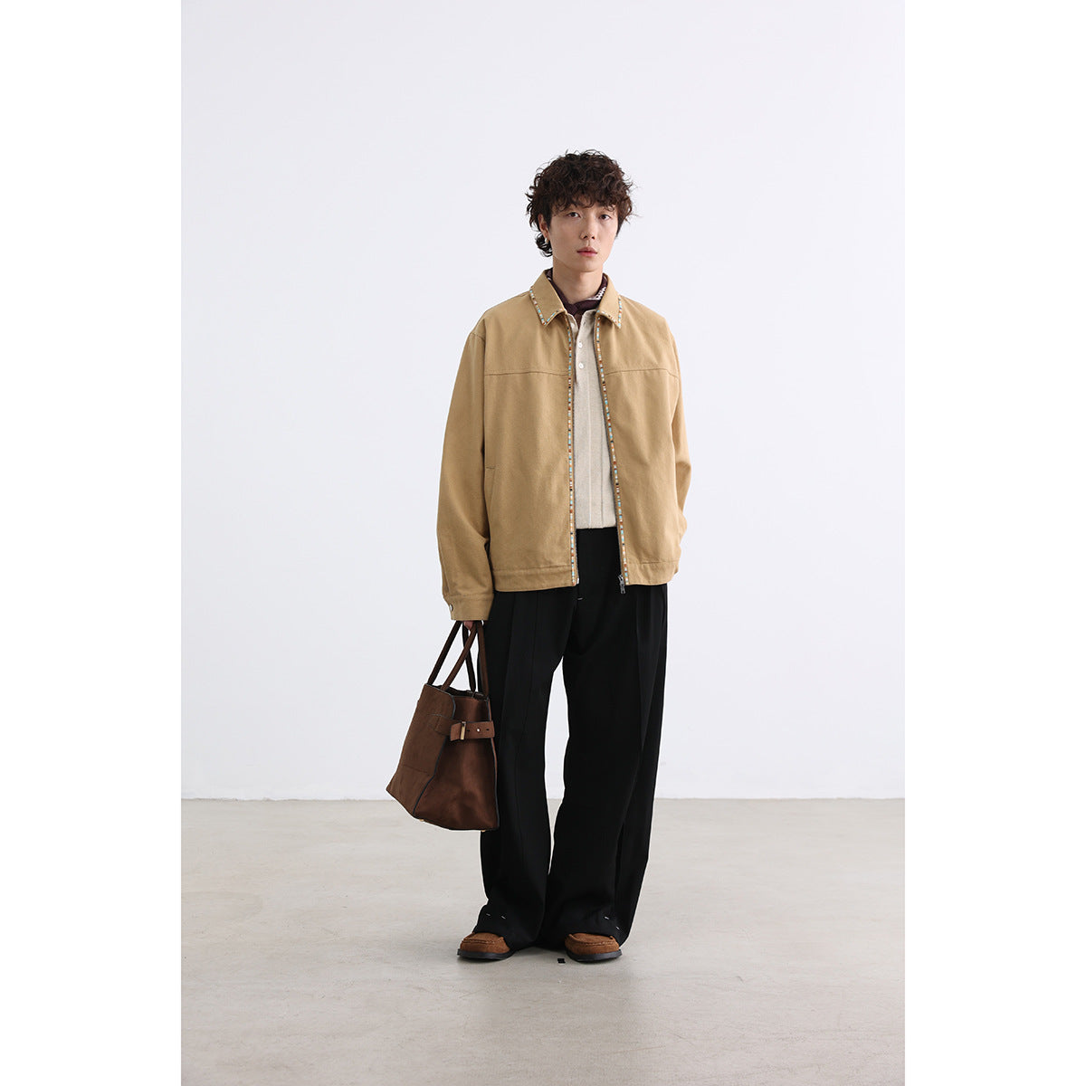 Full-length model wearing a tan jacket with multicolor trim, black trousers, and carrying a brown tote.