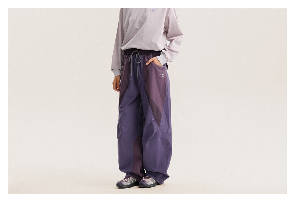 Cropped model wearing loose purple paneled pants with a drawstring waist, shown from a slight front angle.