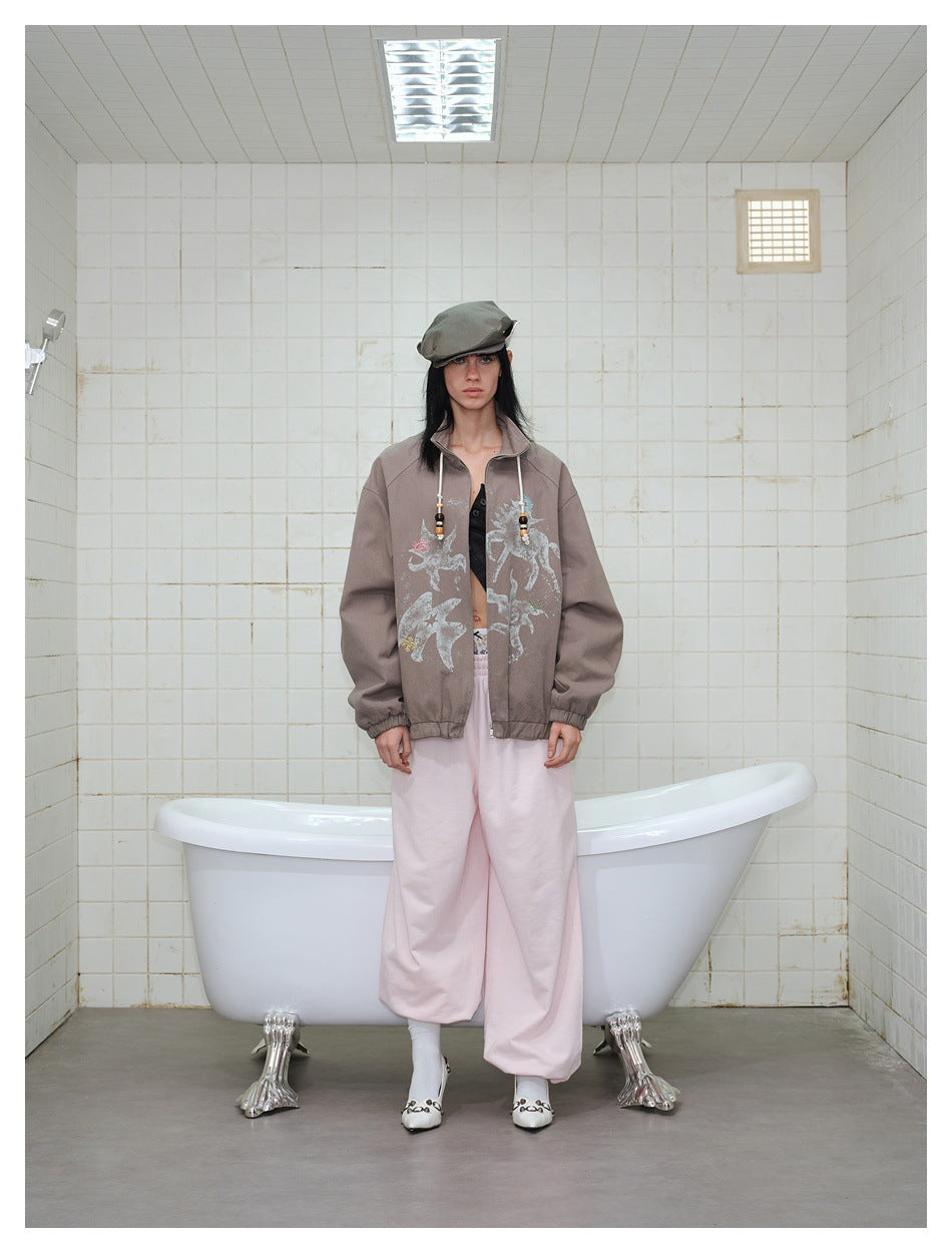 Model wearing a taupe jacket with pale animal and floral graphics, pale pink wide-leg trousers, and a gray cap.