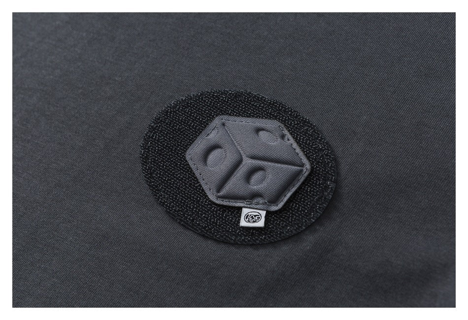 Close-up of a raised dark emblem patch on charcoal T-shirt fabric, with a small symbol tab below.