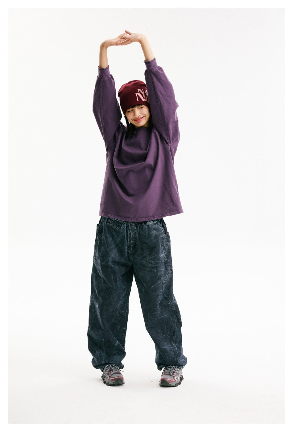 Model stretches overhead in a purple oversized raglan sweatshirt, dark patterned pants, and a burgundy beanie with white