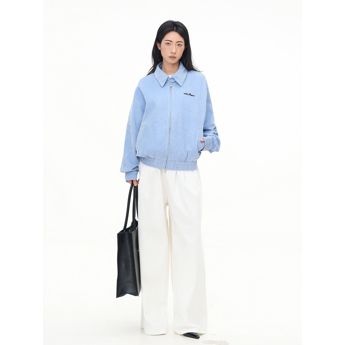 Full-length model wearing a zipped light blue collared jacket and wide-leg white trousers, carrying a black tote.