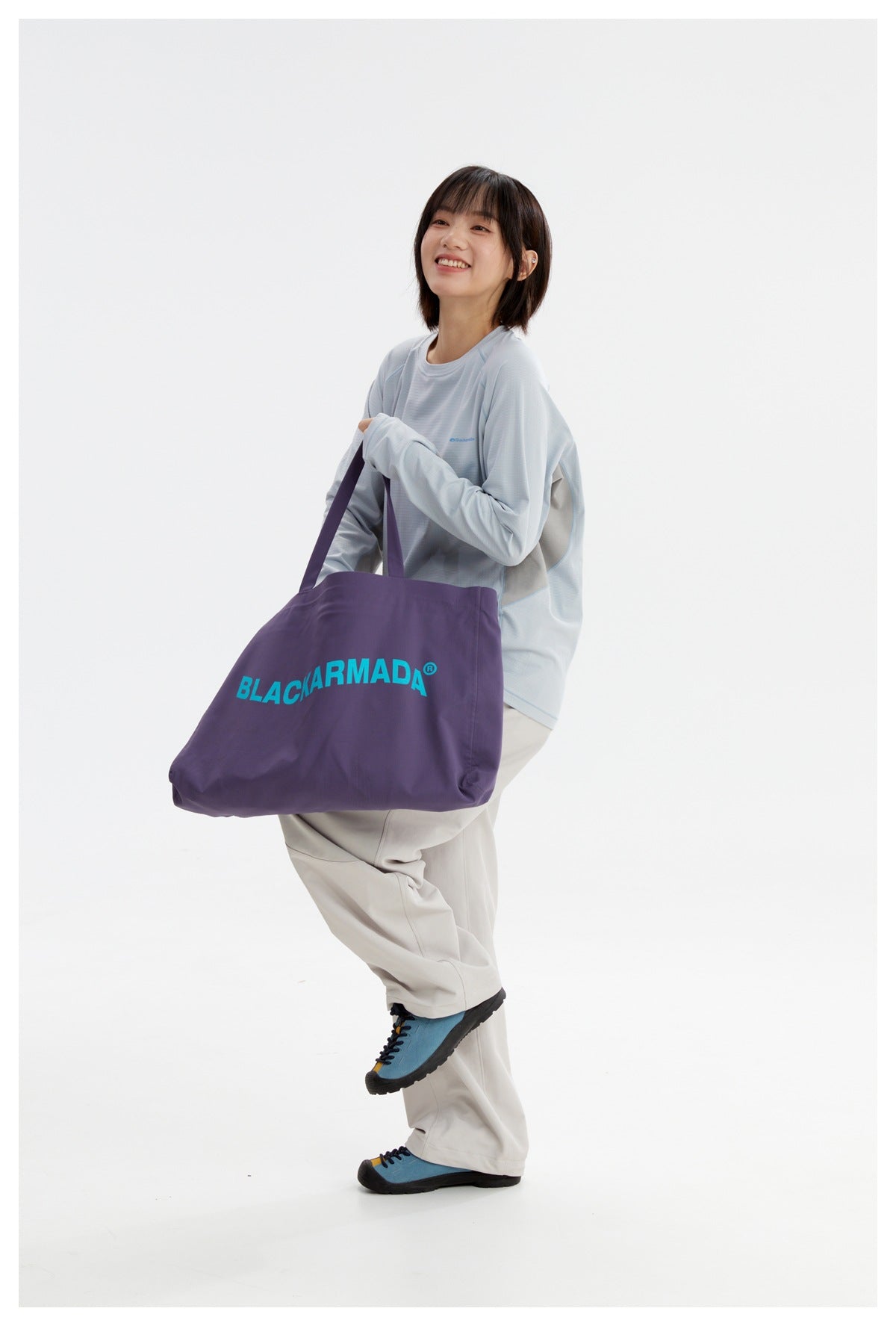 Model wearing a pale blue top and off-white trousers while carrying a large purple tote with turquoise lettering.