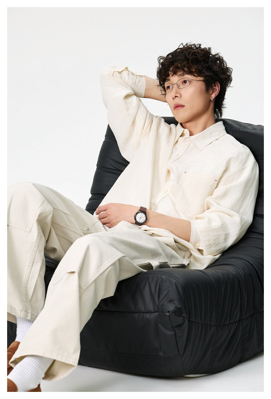 Man seated on a black chair wearing an off-white oversized button-up shirt with chest pocket and cream trousers